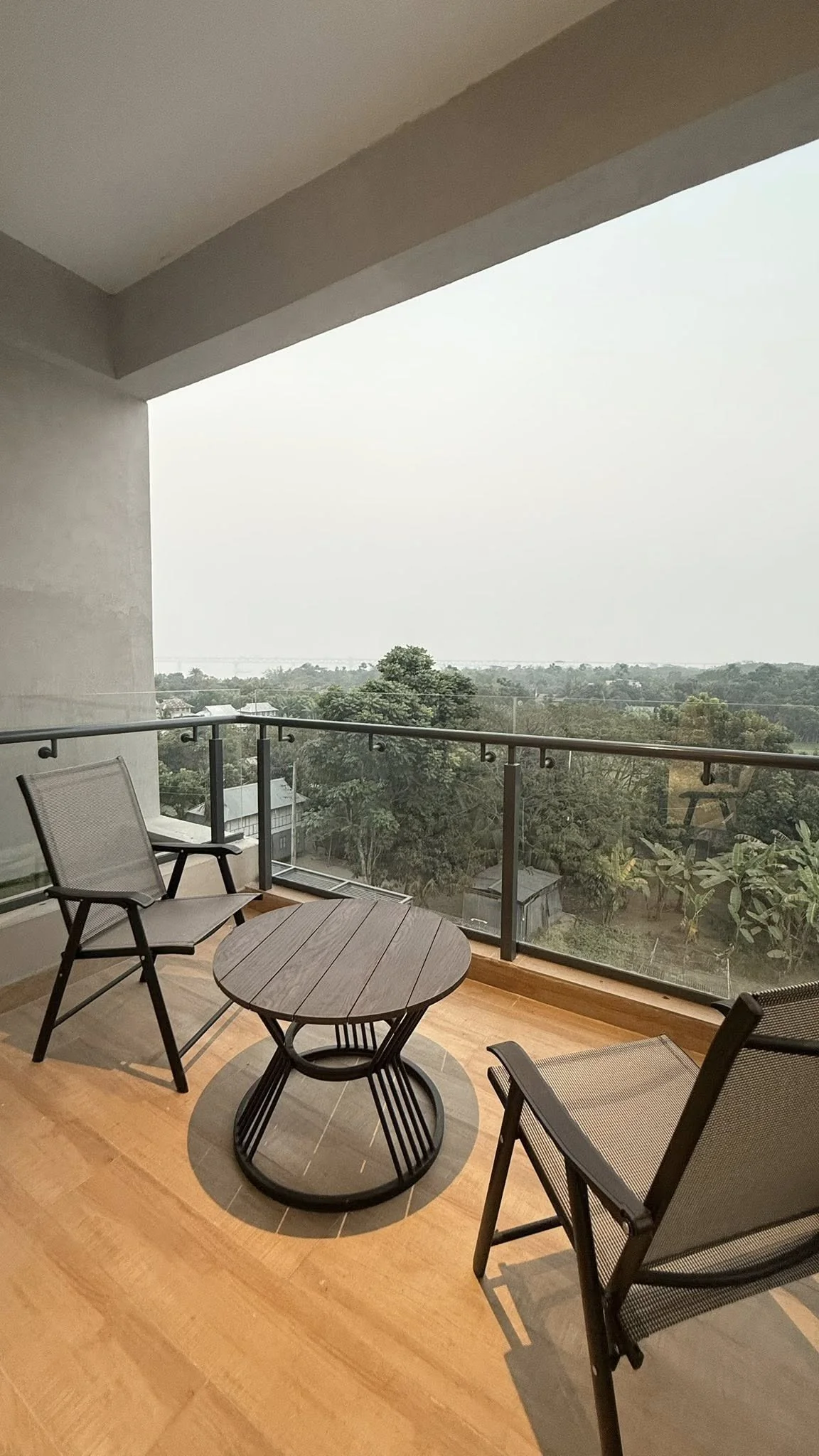 Suite balcony looking out across the surrounding farmland: a small round timber table between two black metal chairs, glass and metal balustrade, treetops in the foreground