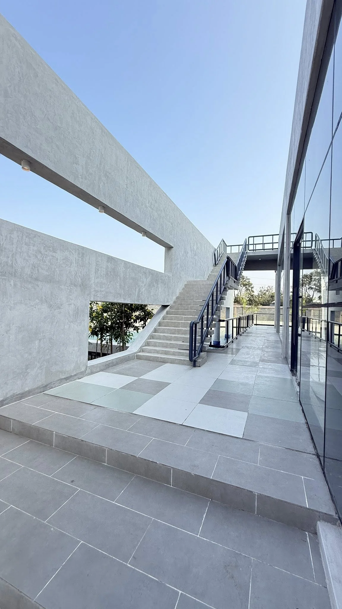External concrete stair connecting the plaza to upper deck level: a tall concrete framing wall with cut openings, blue handrail, glass-clad service block to one side