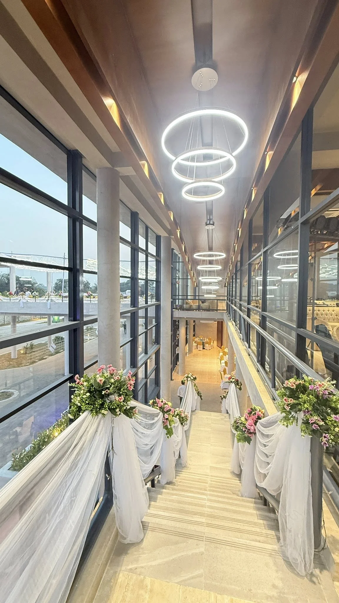 The completed glass-walled corridor between buildings, dressed for an event: tiered ring chandeliers overhead, white drapes and floral bouquets along the railings, the plaza visible through the glazing
