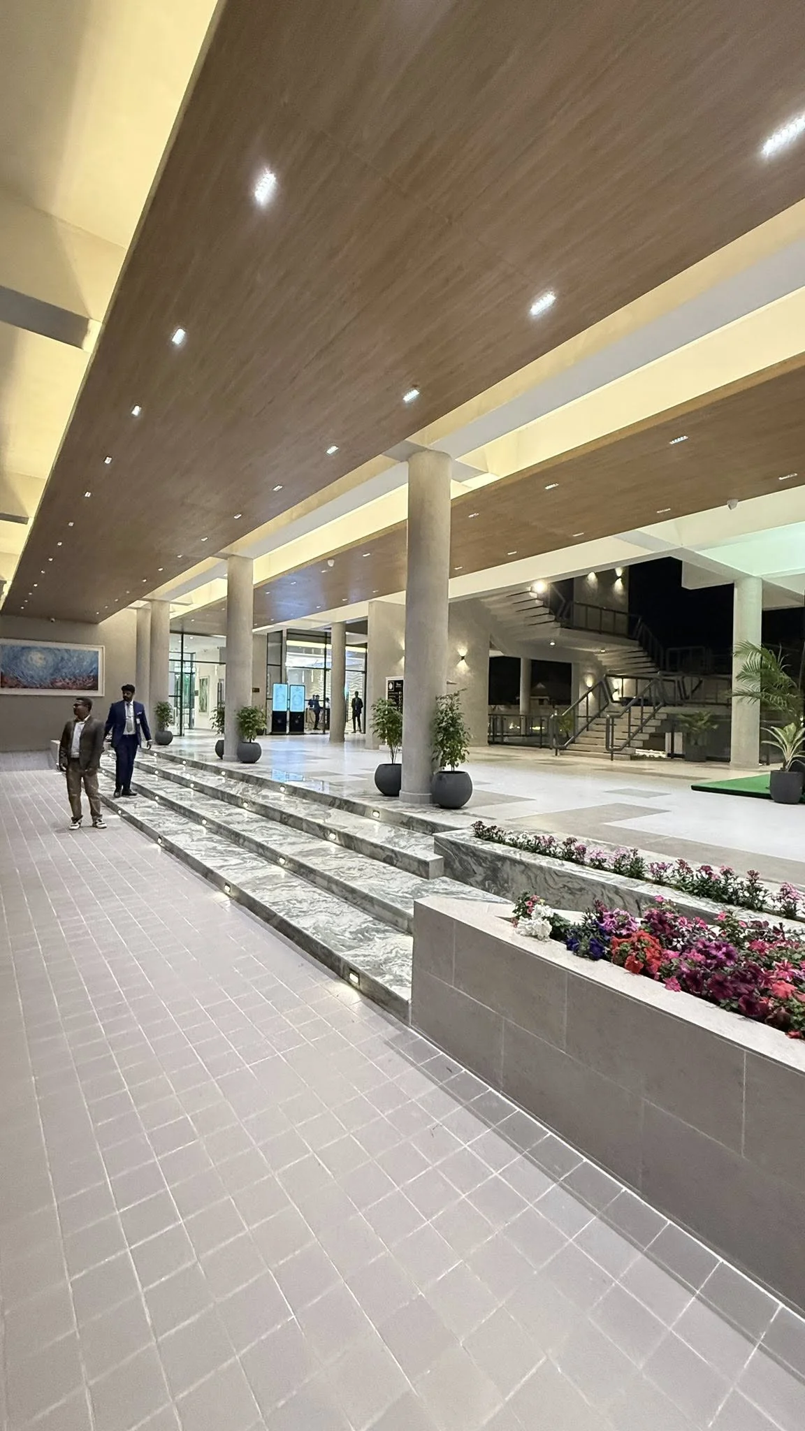 The completed lobby concourse: timber-panelled ceiling, concrete columns lining a long open plate, marble step planters with seasonal flowers, polished tile floor