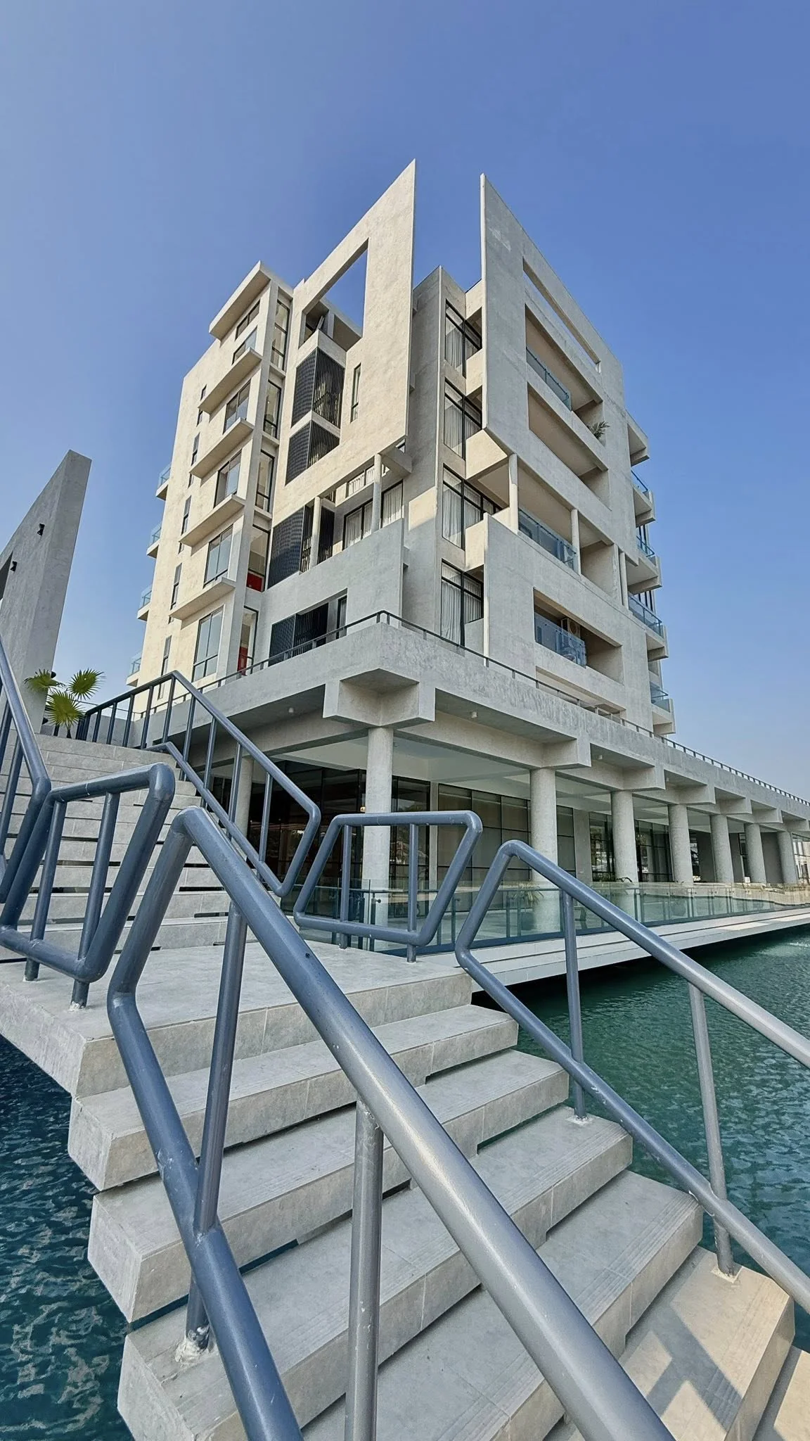 Low-angle view of the completed accommodation block from the water-side, showing the projecting balconies, the plaza colonnade beneath, and the entry stairs across the moat