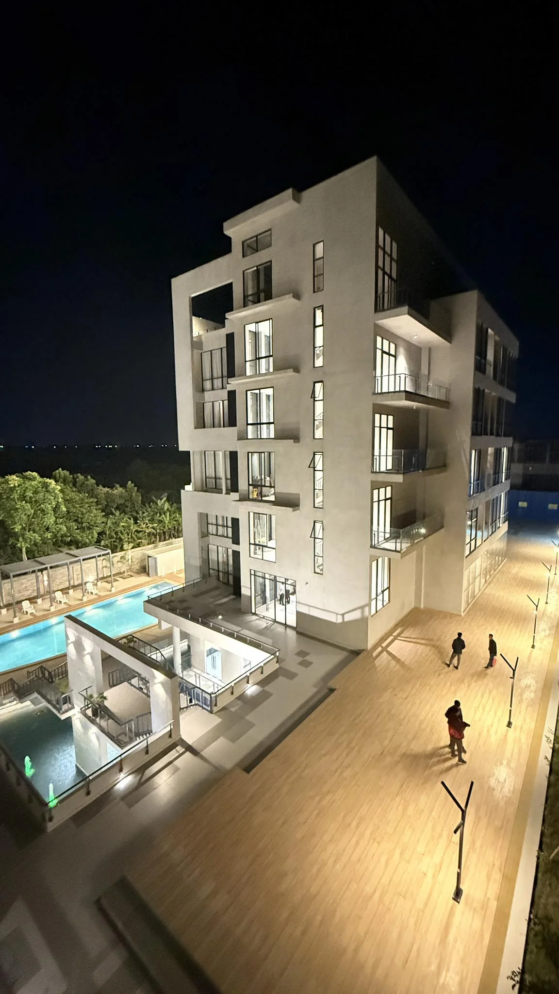 The accommodation block at night, illuminated, with the timber plaza deck below and the swimming pool glowing in the foreground