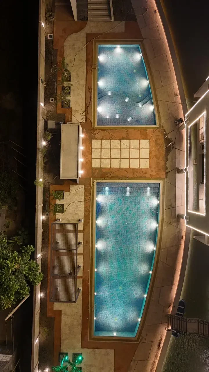 Aerial top-down view of the twin swimming pools at night, lit from below, separated by a paved deck and bordered by the building structure