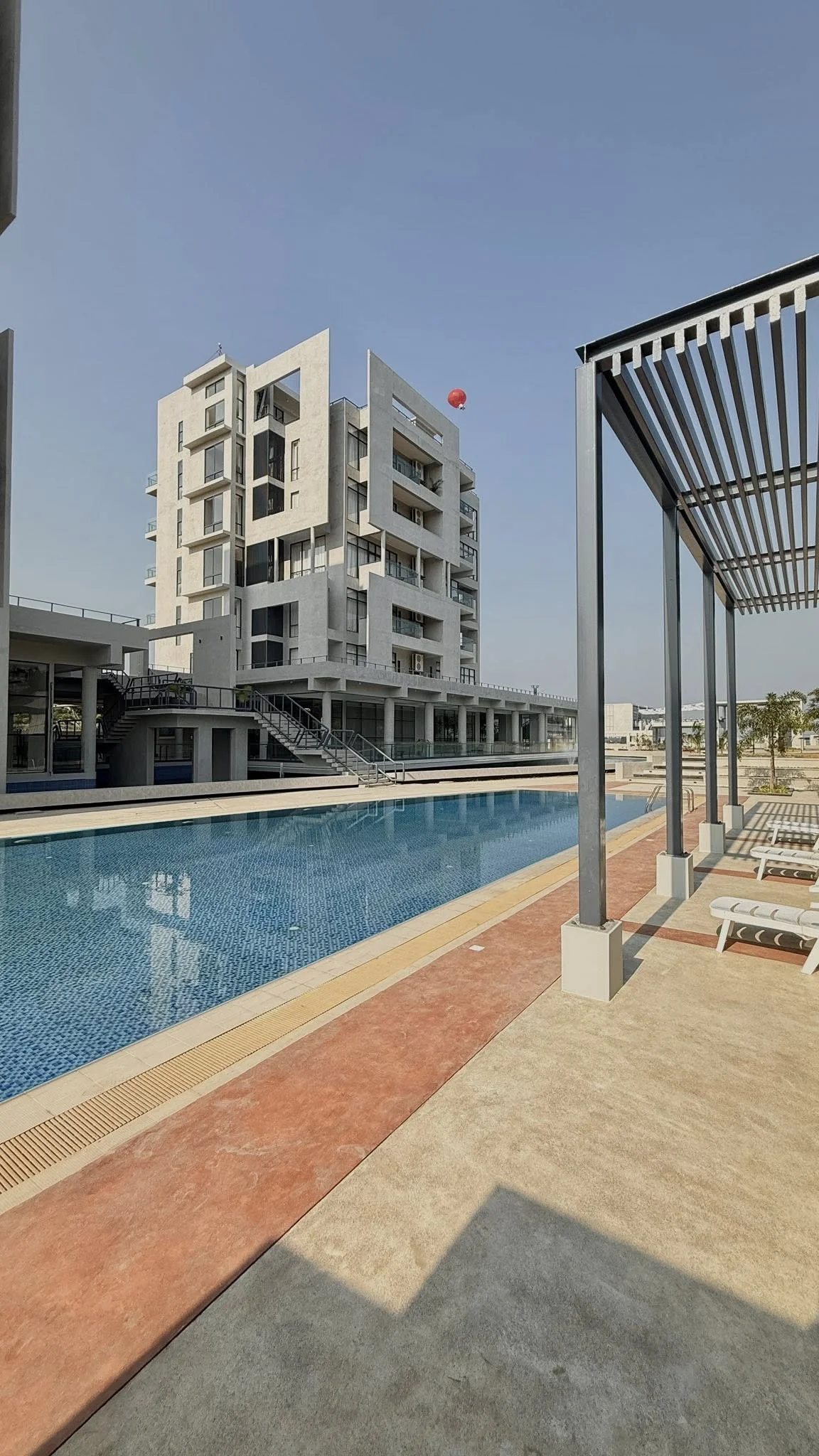 Daytime view of the swimming pool with the steel pergola, the G+5 accommodation block rising behind, sun loungers along the deck, balloon visible in the sky