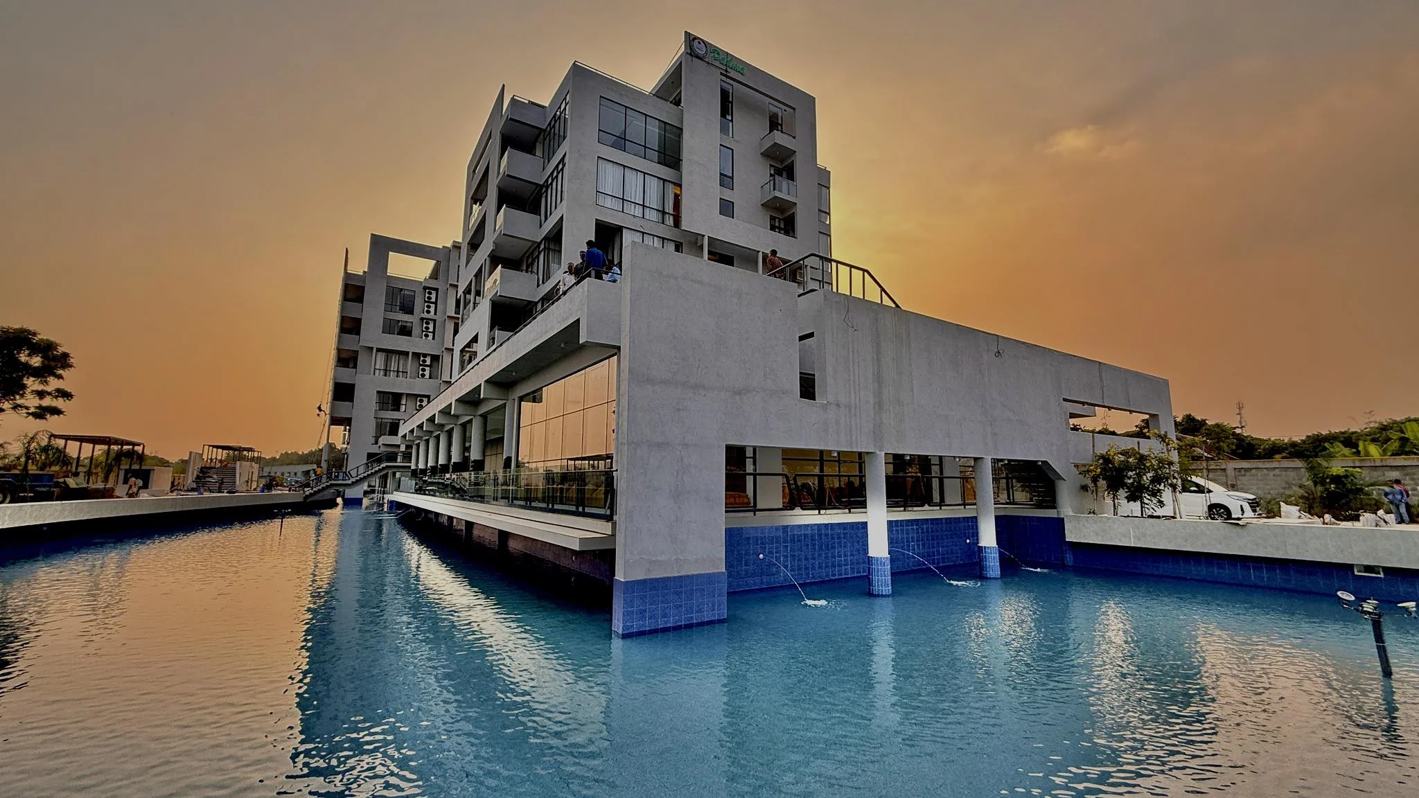 The completed resort at sunset: the G+5 accommodation block rising above the elevated terrace, the swimming pool extending into the foreground reflecting a golden sky, the building's drop-off porch and pool deck visible at plaza level