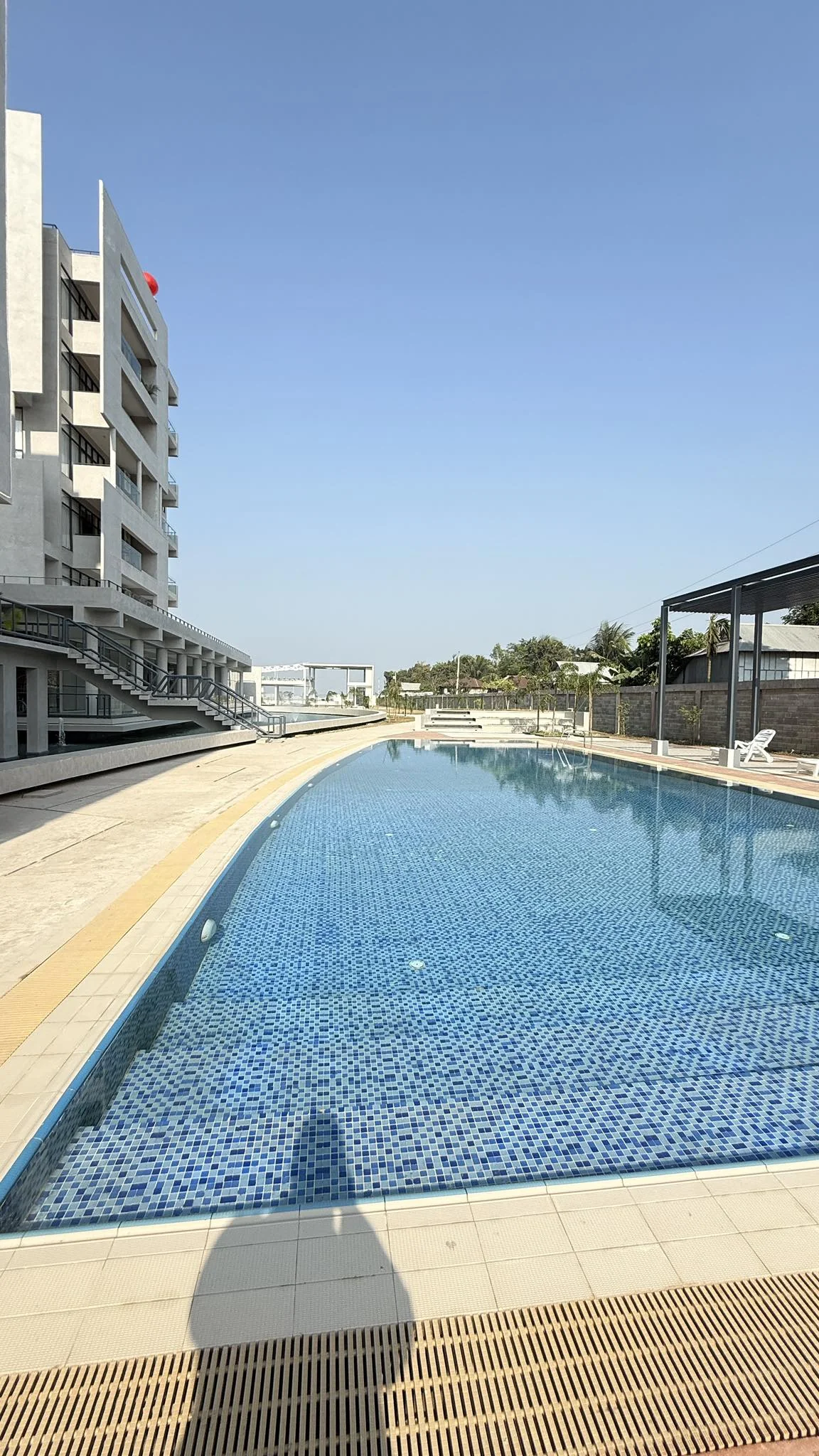 Wide view of the long swimming pool with mosaic-tile finish, the accommodation block on the left, clear sky overhead and the perimeter wall beyond