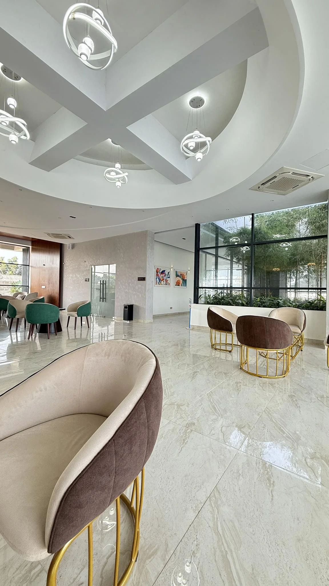 Reception waiting lounge with circular feature ceiling, suspended ring pendant lights, lounge clusters in beige and brown, full-height windows opening to landscaped courtyard