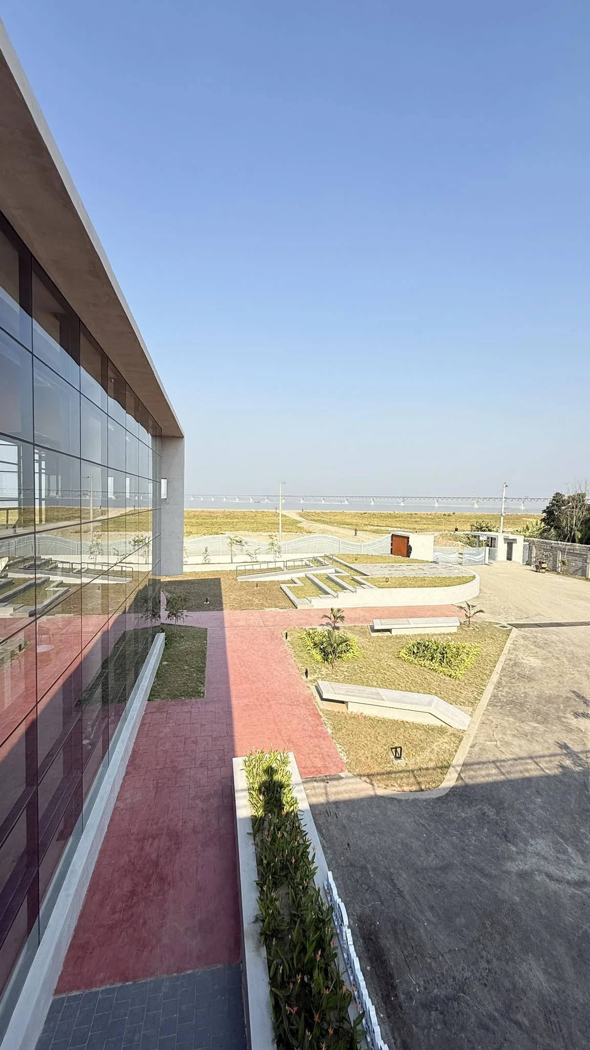 The completed restaurant pavilion exterior: full-height glass facade reflecting the pink walkway and landscape, the Padma River visible beyond at the horizon