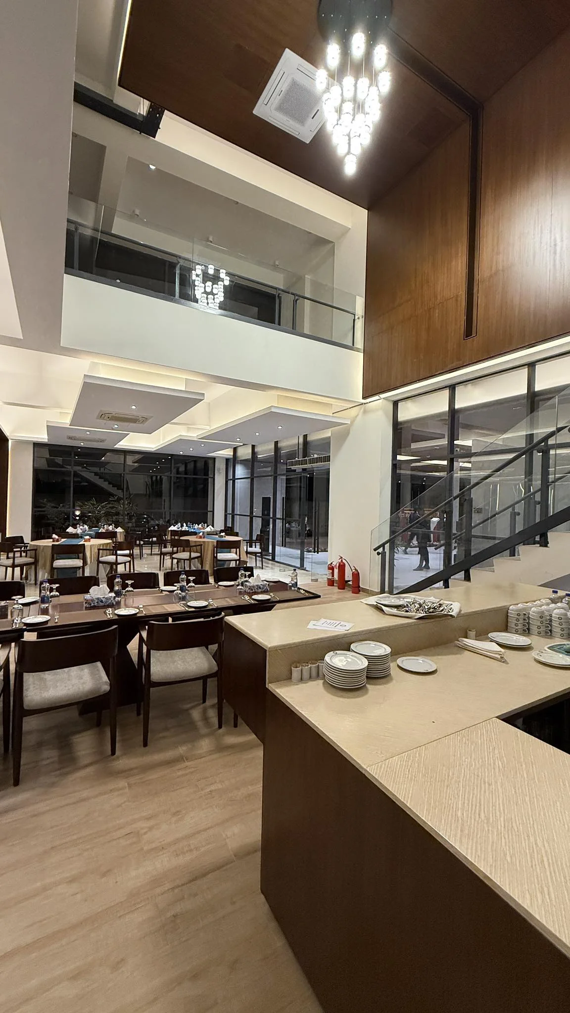 The completed all-day restaurant interior: double-height space, timber-panelled wall, suspended chandelier, dining tables set with linen, glazed mezzanine above
