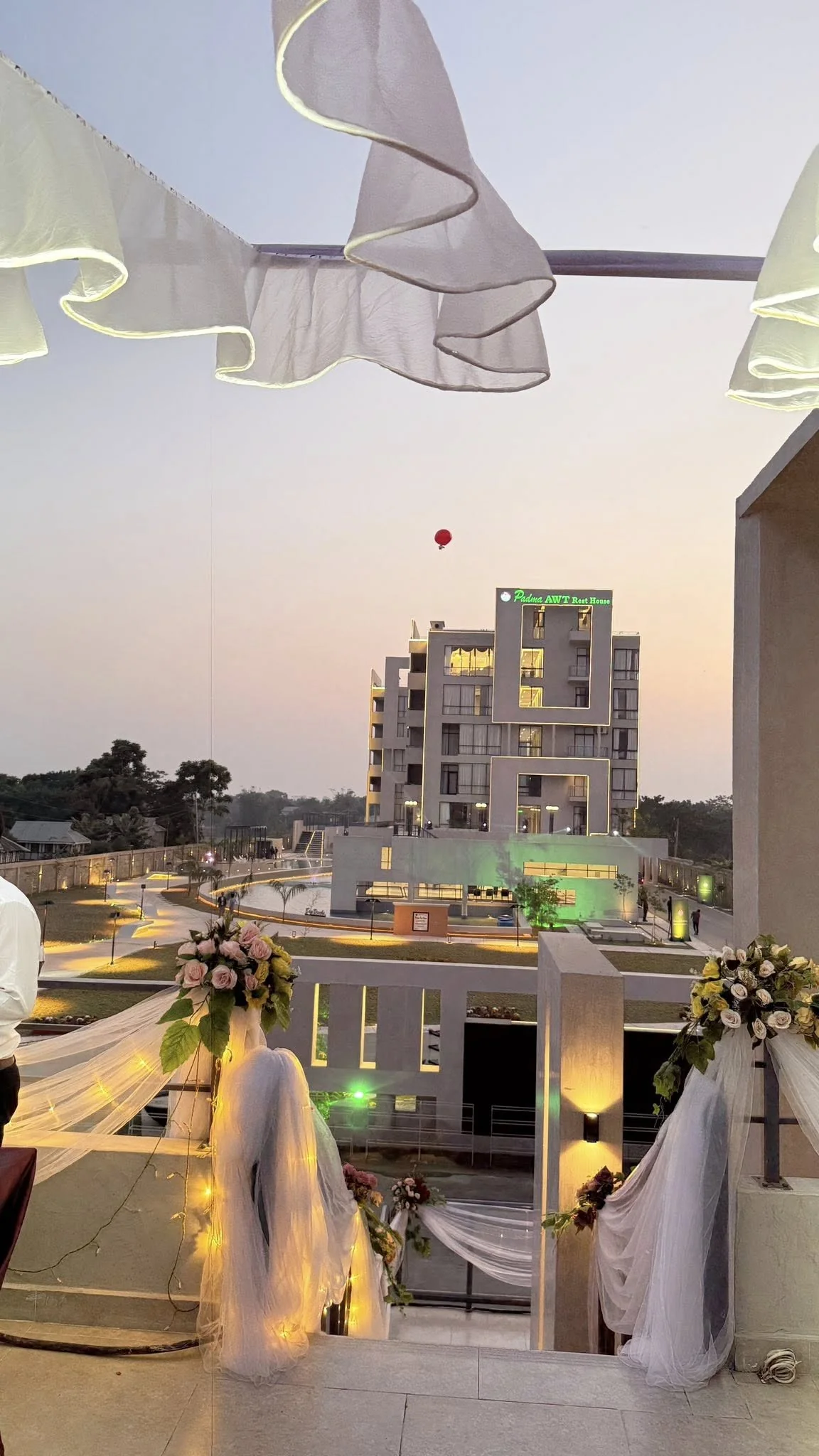The rooftop dressed for the grand opening: floral bouquets and white tulle drapery framing the view across the resort to the lit accommodation block at dusk, hot-air balloon above