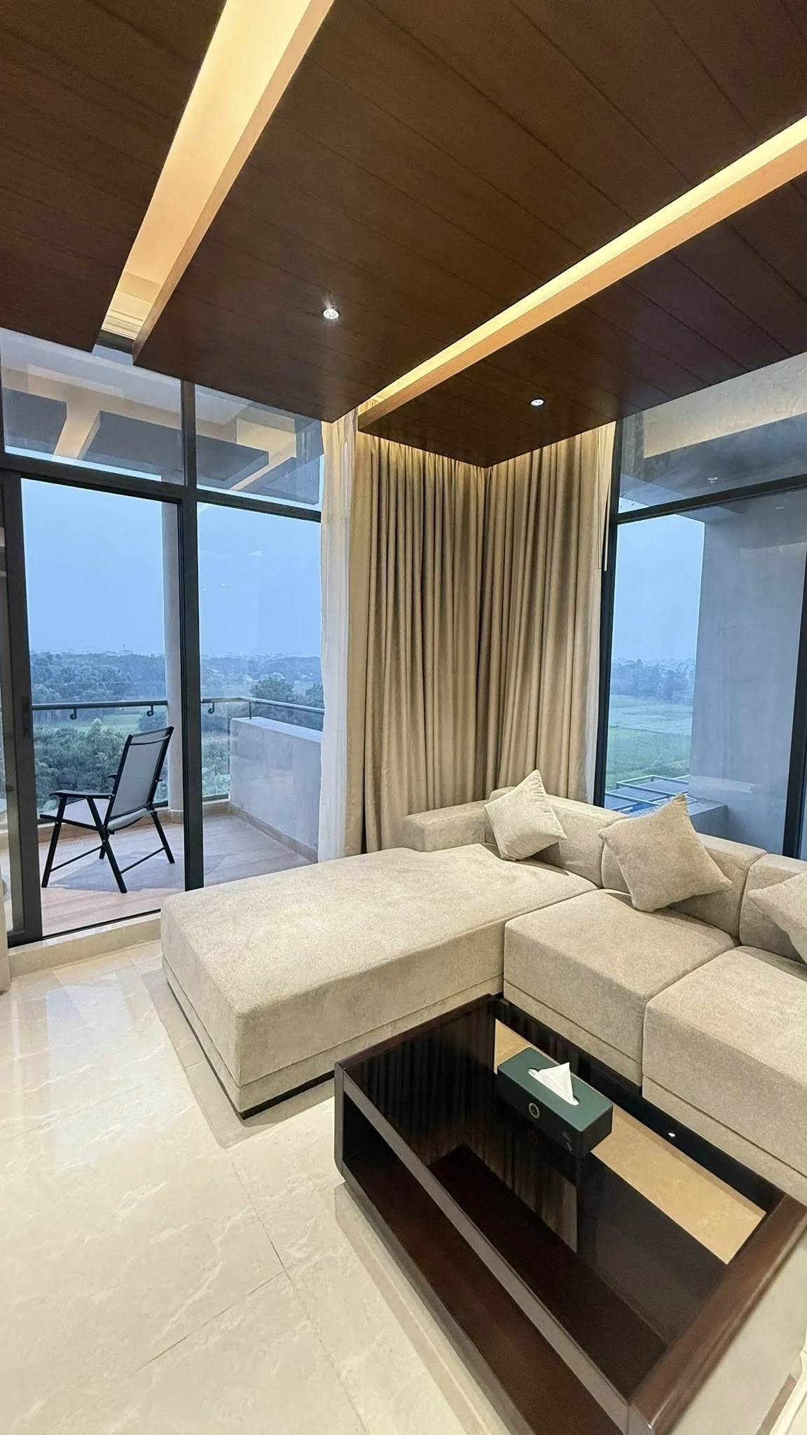Suite seating area with sectional sofa in beige, curtains drawn aside, sliding glass doors opening to a private balcony with a view of the resort grounds and tree canopy