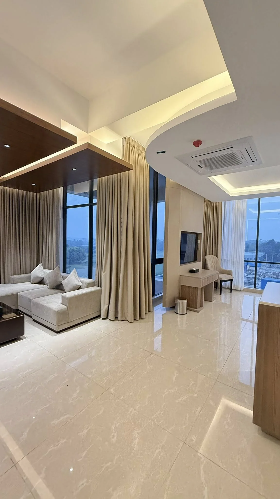 Premium suite living area: long beige sofa against a wood feature wall, full-height curtains drawn open to the river view, polished marble floor, ceiling cove lighting